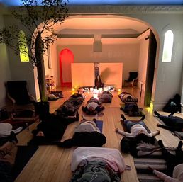 Cozy candlelit sound-bath meditation in a yoga/wellness studio — people lying on mats on a wooden floor while an instructor leads with singing bowls beneath arched alcoves.