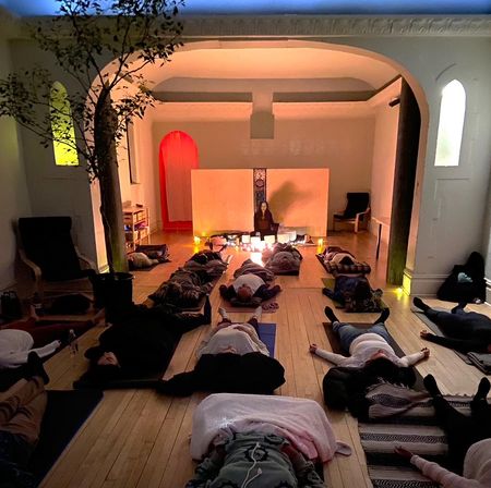 Cozy candlelit sound-bath meditation in a yoga/wellness studio — people lying on mats on a wooden floor while an instructor leads with singing bowls beneath arched alcoves.
