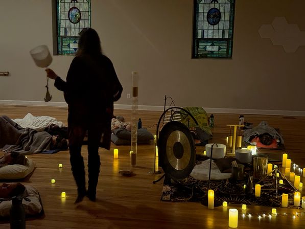 Dim indoor wellness studio group sound bath with a gong, singing bowls, flickering candles and participants resting on mats beneath stained-glass windows