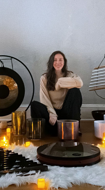 Smiling person seated on the floor in a cozy meditation studio, surrounded by a gong, chimes, crystal singing bowls, mallets and glowing candles for a sound healing session.