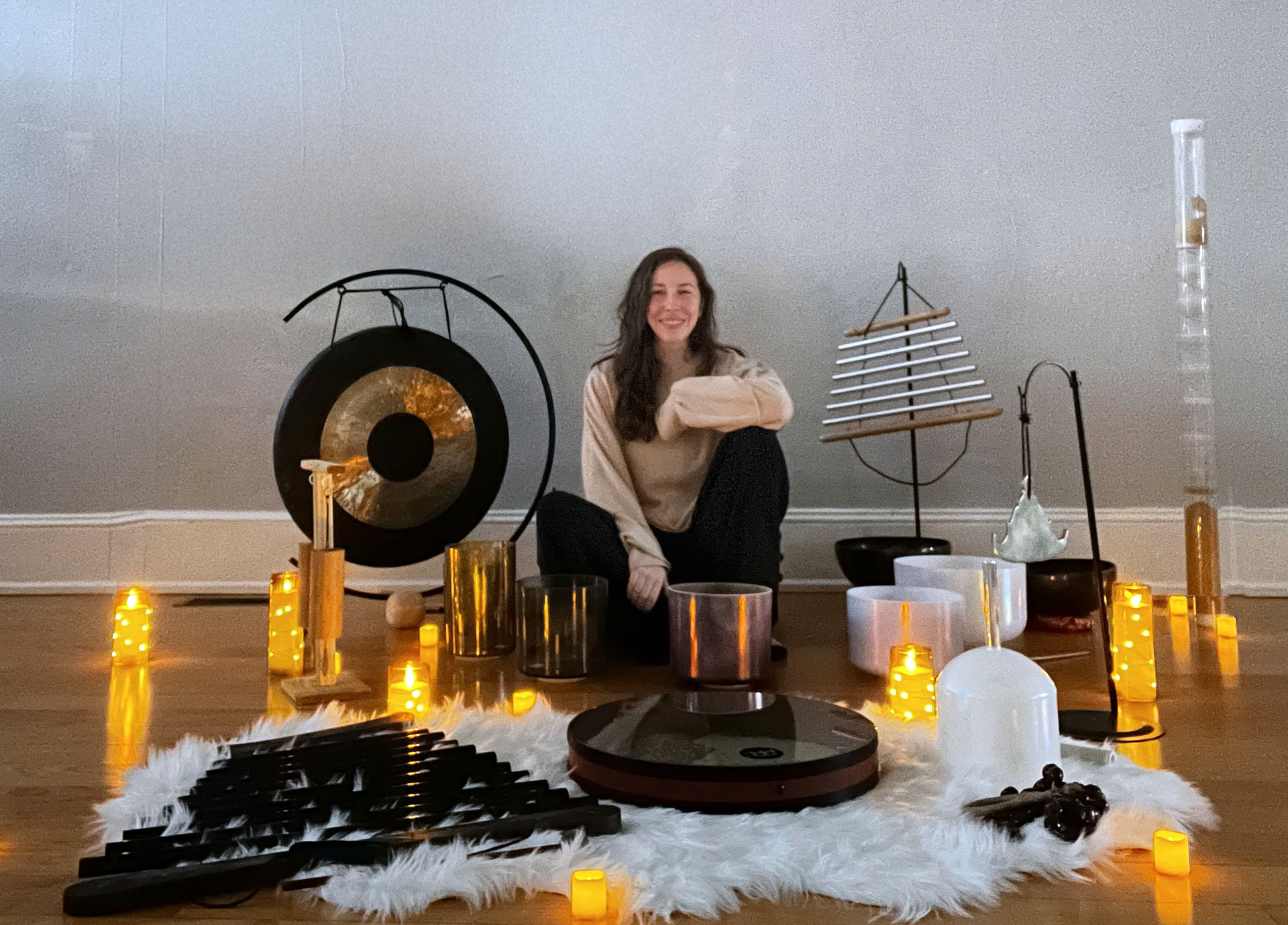 Smiling person seated on the floor in a cozy meditation studio, surrounded by a gong, chimes, crystal singing bowls, mallets and glowing candles for a sound healing session.