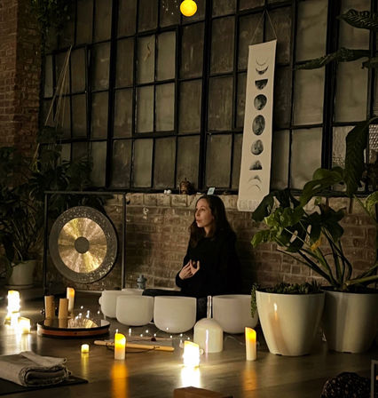 Cozy candlelit sound bath in an urban loft studio — a person meditating among crystal singing bowls, a gong, potted plants and a moon-phase tapestry against an exposed brick wall.