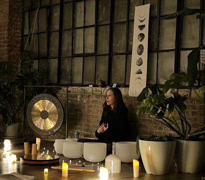 Cozy candlelit sound bath in an urban loft studio — a person meditating among crystal singing bowls, a gong, potted plants and a moon-phase tapestry against an exposed brick wall.