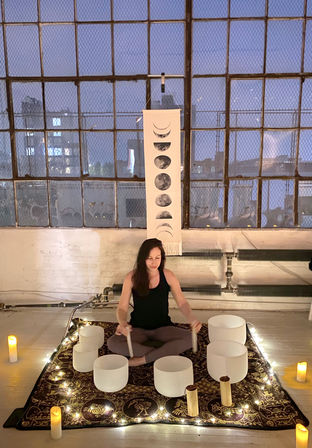 Person seated cross-legged in an urban loft hosting a candlelit sound bath with crystal singing bowls on a patterned rug, string lights, moon-phase wall hanging, and city skyline beyond large industrial paned windows.