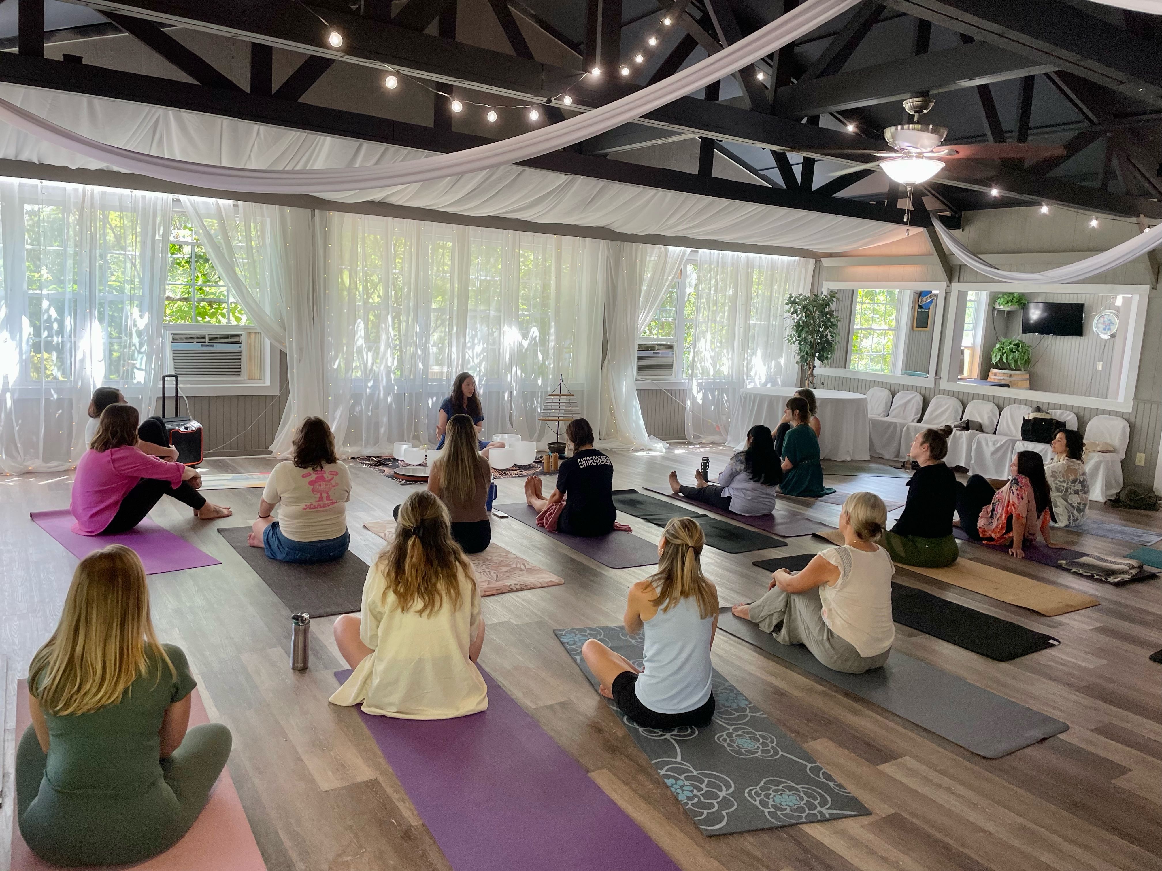 Community yoga and meditation class in a bright, airy studio with exposed dark beams, white drapery and string lights; participants seated on colorful mats facing an instructor by sheer sunlit windows.