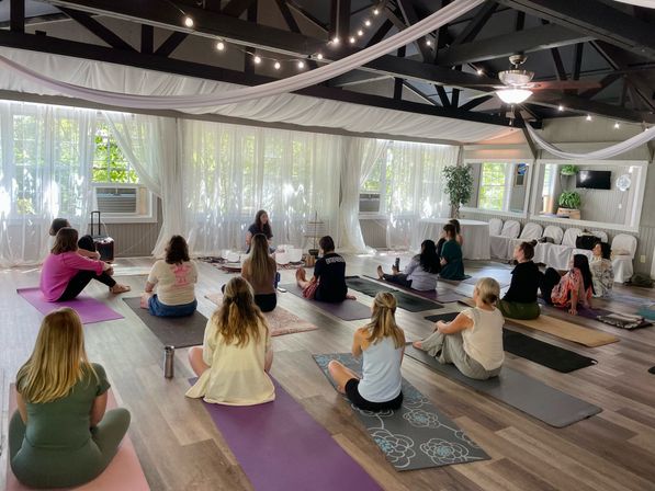 Community yoga and meditation class in a bright, airy studio with exposed dark beams, white drapery and string lights; participants seated on colorful mats facing an instructor by sheer sunlit windows.