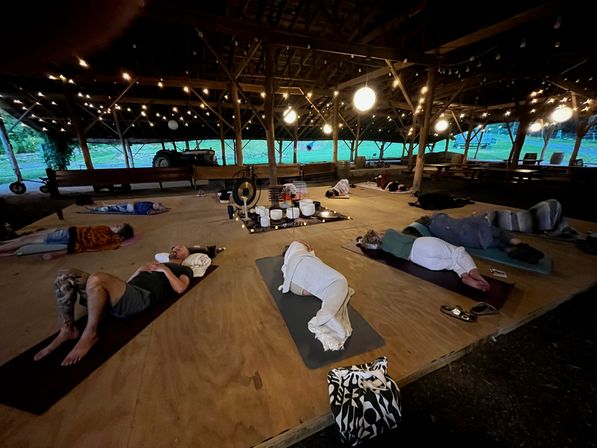 Group reclining on yoga mats for a sound-bath meditation inside a rustic open-sided farm barn pavilion at dusk, with hanging string lights, gongs and singing bowls on a central wooden platform.