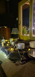 Cozy living-room sound bath setup at night: crystal singing bowls, metal bowls, hanging chimes and a triangular tuning-frame on a celestial cloth, surrounded by LED candles and string lights beside glass patio doors with a potted fern outside.