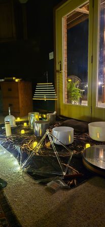 Cozy living-room sound bath setup at night: crystal singing bowls, metal bowls, hanging chimes and a triangular tuning-frame on a celestial cloth, surrounded by LED candles and string lights beside glass patio doors with a potted fern outside.