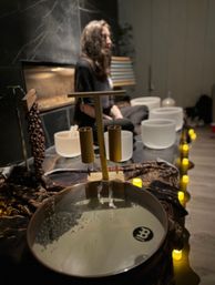 Person seated behind frosted crystal singing bowls and a large metal sound bowl with mallets and glowing candles in a dim wellness studio during a relaxing sound bath meditation session.