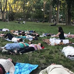 Group sound bath in an urban park: people lying on blankets on a grassy lawn while a practitioner plays crystal singing bowls beneath tall trees at dusk.