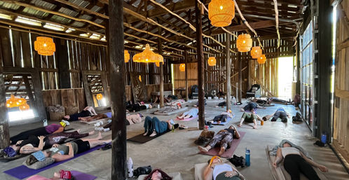 Group yoga/relaxation session in a rustic wooden barn studio — participants lying in Savasana on mats beneath warm woven pendant lights and open-beam ceiling, sunlit countryside retreat vibe.
