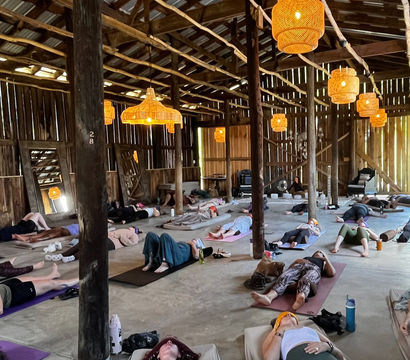 Group yoga/relaxation session in a rustic wooden barn studio — participants lying in Savasana on mats beneath warm woven pendant lights and open-beam ceiling, sunlit countryside retreat vibe.