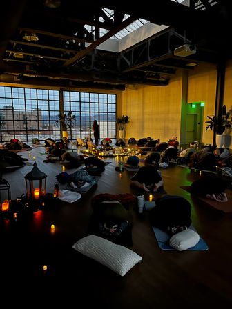 Candlelit restorative yoga class in an urban loft studio at dusk — students in child’s pose on mats around a glowing circle of candles and string lights, large industrial windows framing a city skyline.