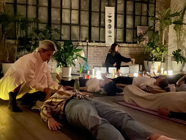 Group sound bath in a cozy loft — participants lie on mats amid candles, potted plants and crystal singing bowls while a facilitator plays under a moon-phase banner.