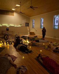 Candlelit sound bath in a warm wood-ceiling meditation hall with stained-glass windows; participants reclining on mats and blankets around a gong and instruments.