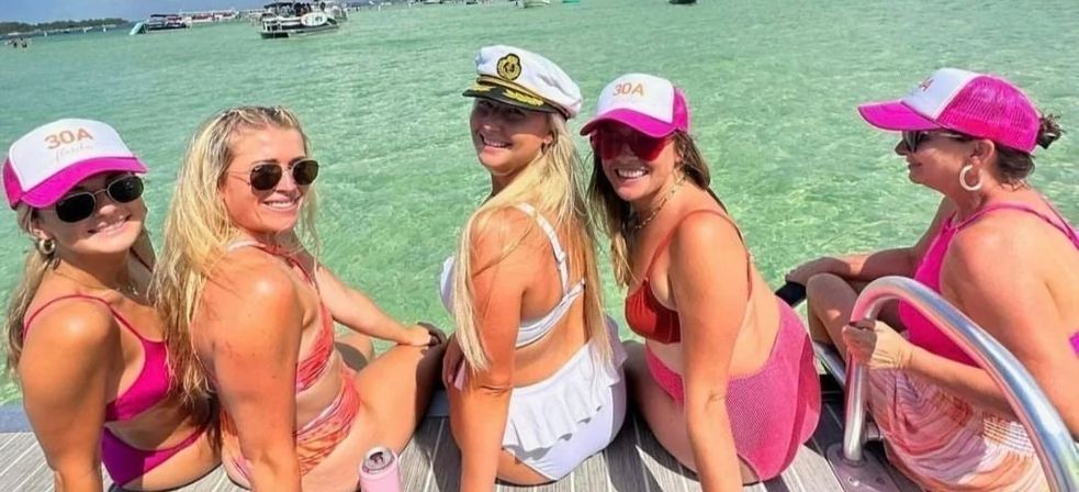 Five women in colorful swimsuits and pink caps sitting on a dock, one wearing a captain's hat, smiling at the camera with clear turquoise water and boats nearby — sunny summer boating scene.