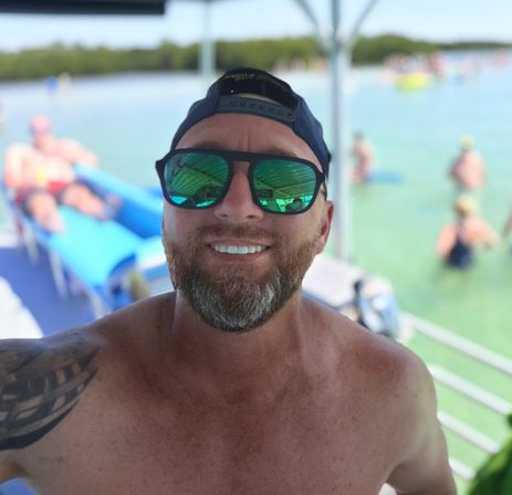 Smiling shirtless man in a backwards cap and mirrored sunglasses taking a boat selfie over clear turquoise water with swimmers and floating loungers in the background.