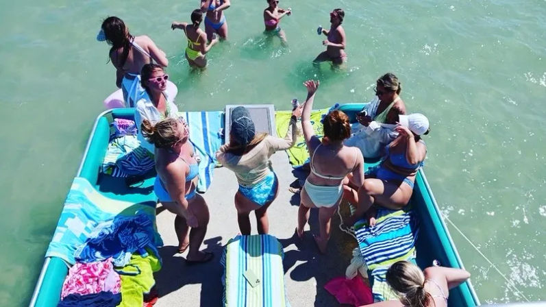 Overhead view of a summer outing on a teal pontoon boat with a group of friends in colorful swimwear and towels, some dancing on the deck while others wade in shallow turquoise water.