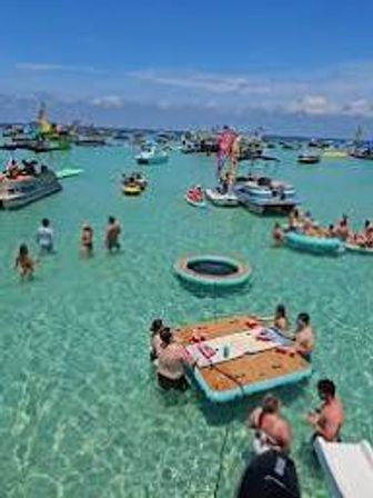 People lounging on floating platforms, inner tubes and anchored boats in a shallow turquoise tropical bay on a sunny blue-sky day