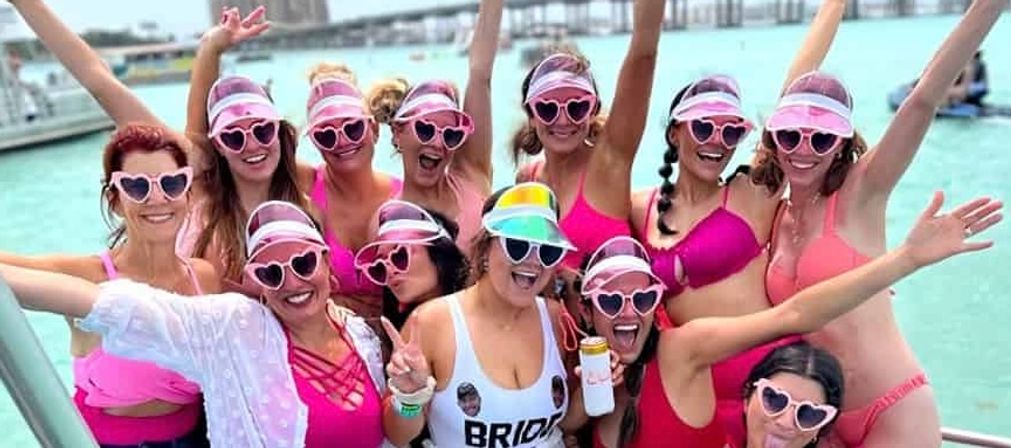 Bachelorette boat party with a cheering group of women in pink swimsuits, heart-shaped sunglasses and visors on a sunny day over turquoise coastal water and marina in the background.