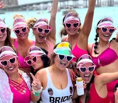 Bachelorette boat party with a cheering group of women in pink swimsuits, heart-shaped sunglasses and visors on a sunny day over turquoise coastal water and marina in the background.