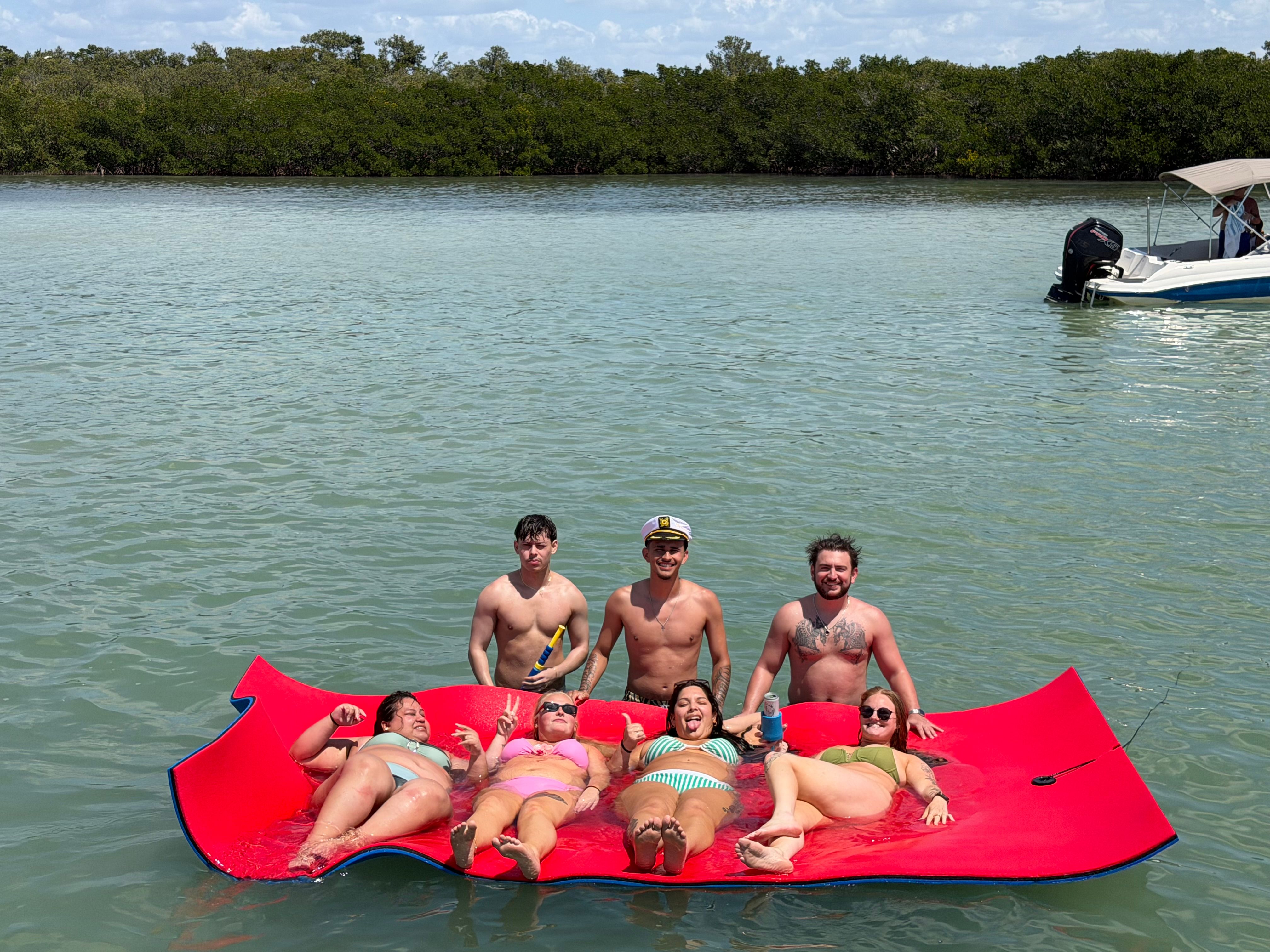 Seven adults enjoying a sunny day on a large red floating mat in a calm blue‑green coastal inlet with mangrove shoreline; three shirtless men stand behind while four people in colorful swimsuits recline, a small motorboat visible in the background.