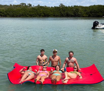 Seven adults enjoying a sunny day on a large red floating mat in a calm blue‑green coastal inlet with mangrove shoreline; three shirtless men stand behind while four people in colorful swimsuits recline, a small motorboat visible in the background.