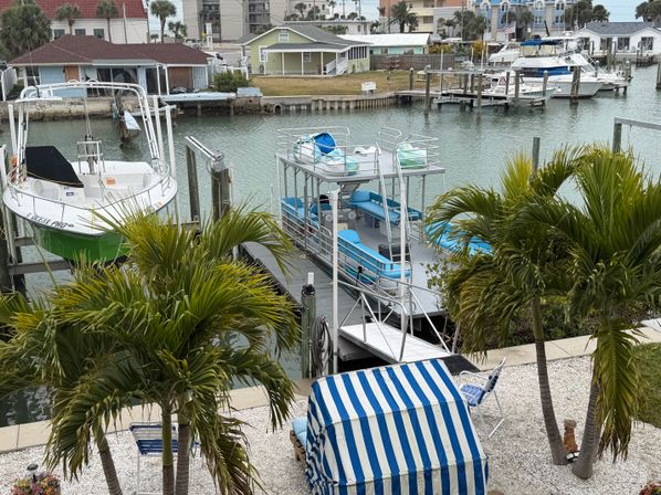 Tropical waterfront canal with palm trees and turquoise water, a blue-and-white striped beach cabana, and docked pontoon and powerboats at residential docks.