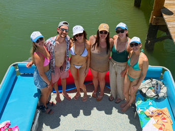 Six friends in swimwear and caps smiling on a bright blue pontoon boat beside a wooden dock over green lake water on a sunny summer day