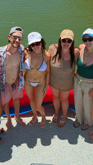 Six friends in swimwear and caps smiling on a bright blue pontoon boat beside a wooden dock over green lake water on a sunny summer day