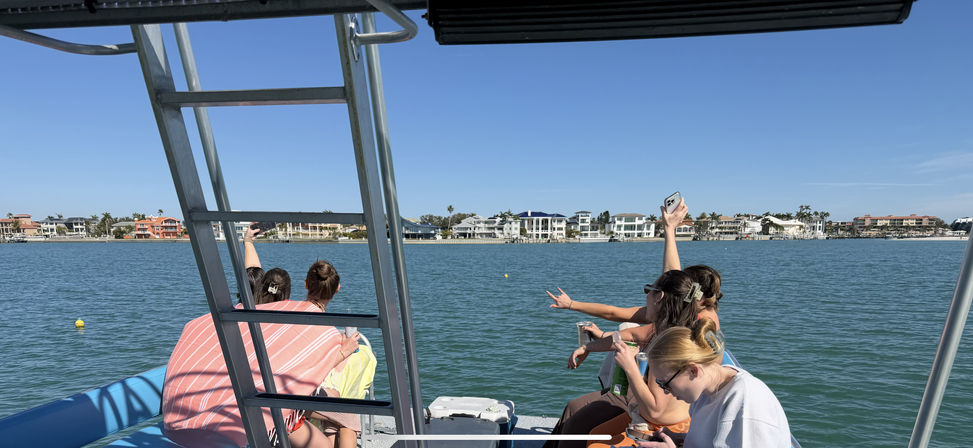Group of friends on a pontoon boat taking selfies and sipping drinks under a clear blue sky, cruising a residential coastal bay lined with waterfront homes