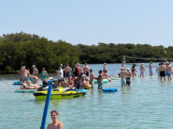 Crowded sandbar party in shallow turquoise water by mangroves, people on inflatables and a beach-volleyball net, plus a bright yellow jet ski under a clear blue sky.