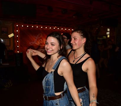 Two smiling friends dancing cheek-to-cheek on a dim bar dance floor, wearing denim outfits with warm marquee bulbs and wooden stage backdrop.