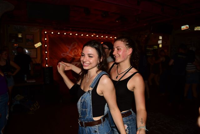 Two smiling friends dancing cheek-to-cheek on a dim bar dance floor, wearing denim outfits with warm marquee bulbs and wooden stage backdrop.