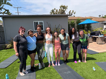 Eight women in activewear smiling and posing on yoga mats in a sunny suburban backyard after an outdoor workout, with artificial turf, palm plants and a blue patio umbrella in the background.