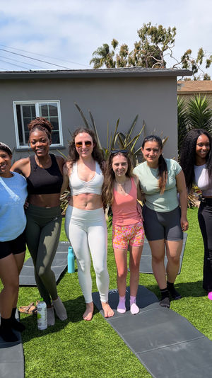 Eight women in activewear smiling and posing on yoga mats in a sunny suburban backyard after an outdoor workout, with artificial turf, palm plants and a blue patio umbrella in the background.