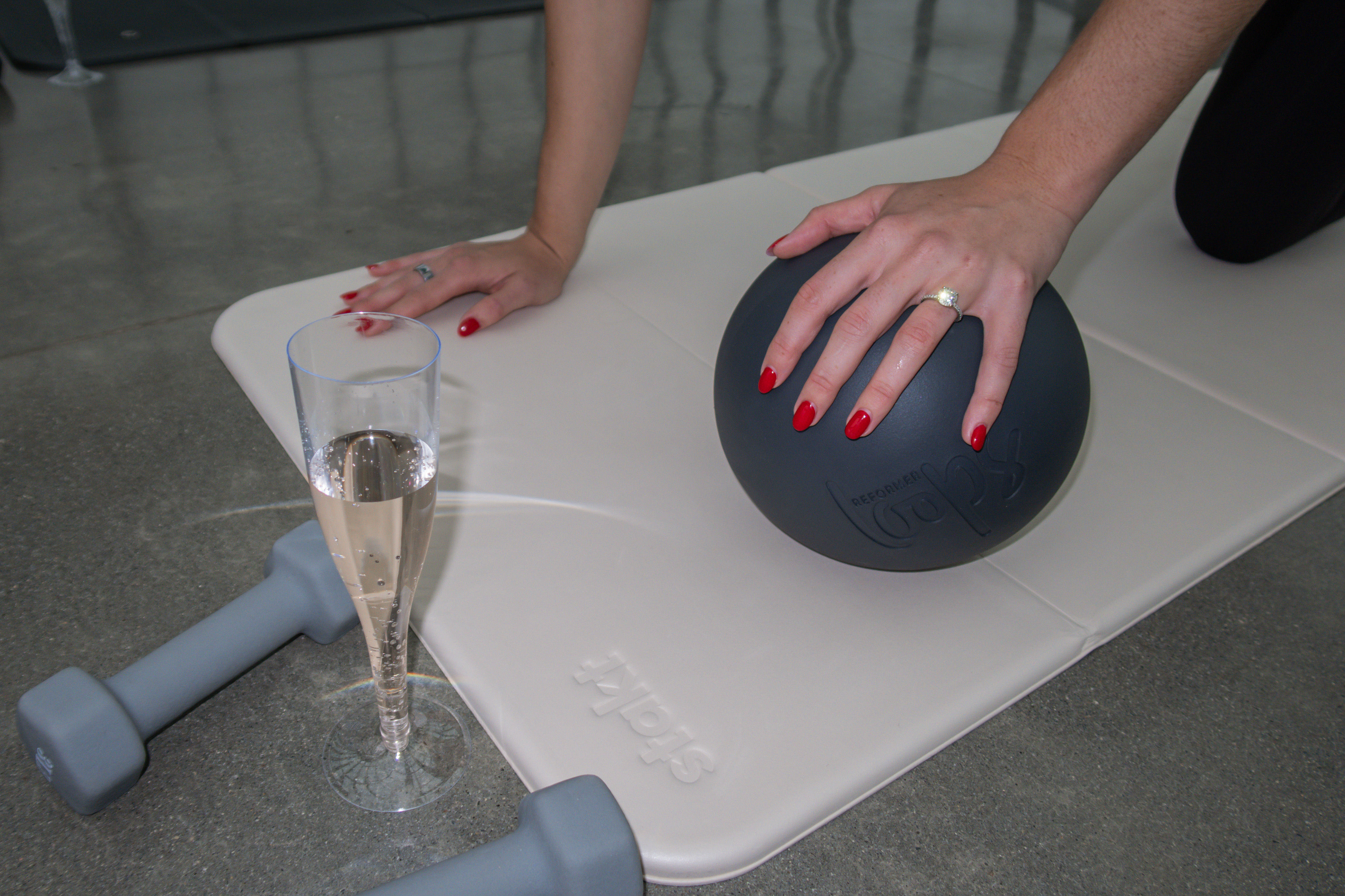 Hands with red nails pressing a black medicine ball on a light exercise mat, with gray dumbbells and a champagne flute on a concrete floor.