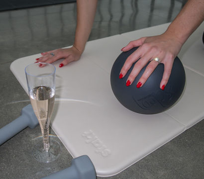 Hands with red nails pressing a black medicine ball on a light exercise mat, with gray dumbbells and a champagne flute on a concrete floor.