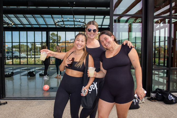 Three smiling women in black activewear holding drinks on a sunny patio outside a modern glass‑walled fitness studio, with yoga mats and workout bags visible inside — post‑workout vibes.