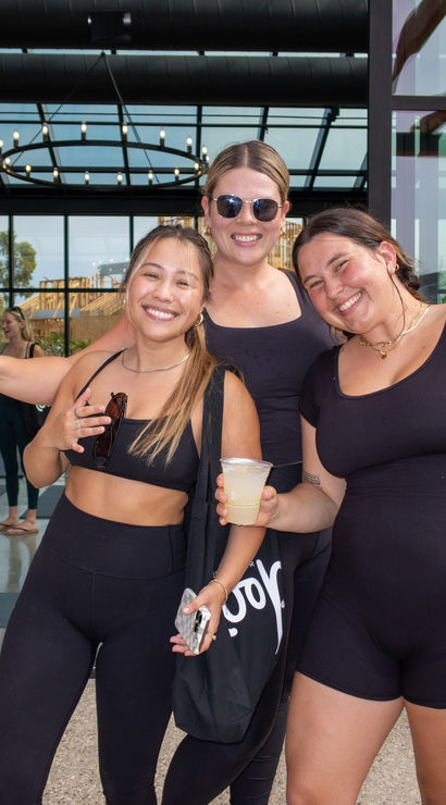 Three smiling women in black activewear holding drinks on a sunny patio outside a modern glass‑walled fitness studio, with yoga mats and workout bags visible inside — post‑workout vibes.