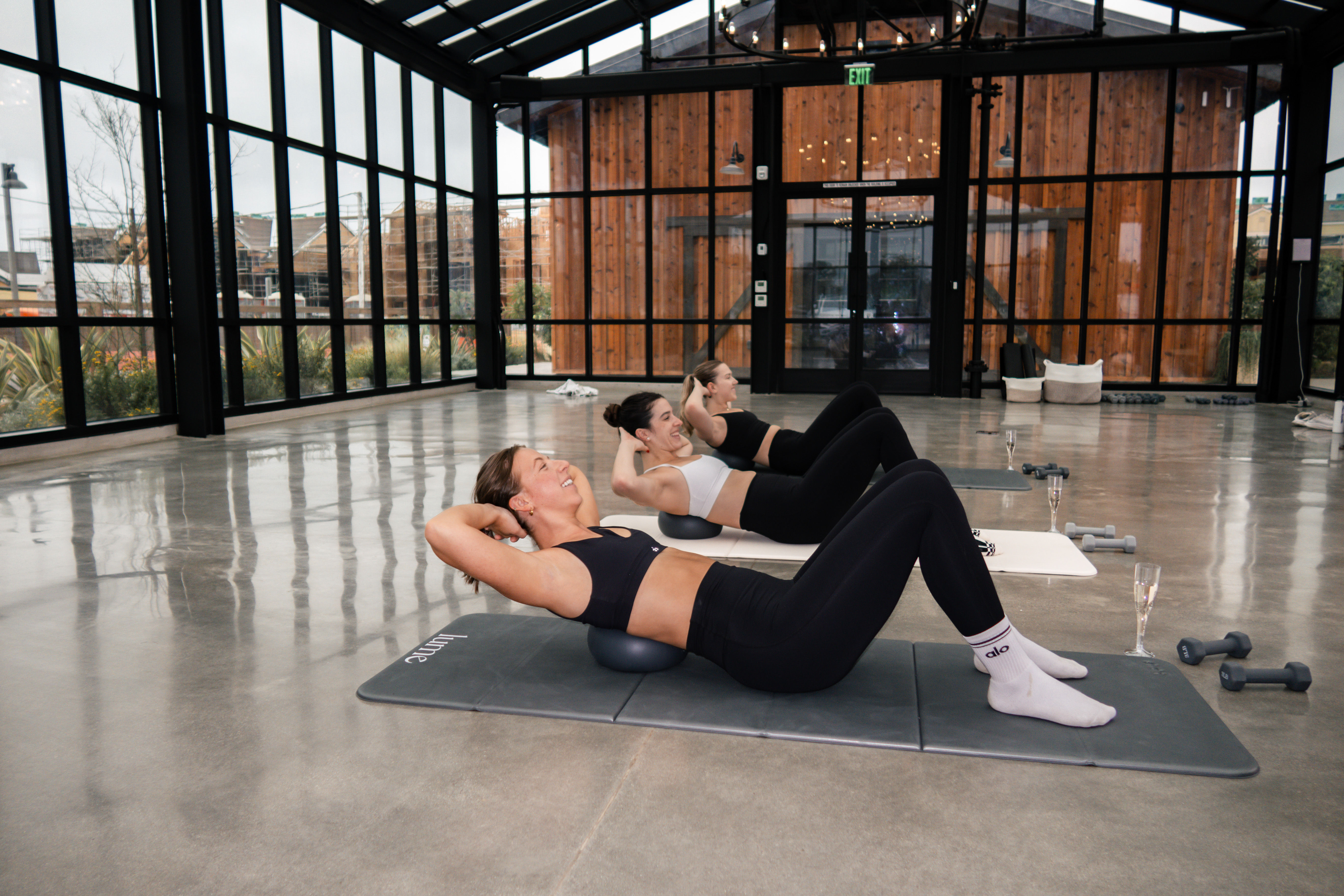 Three women in athletic wear doing core crunches on small stability balls in a bright glass-walled fitness studio with concrete floor, mats, dumbbells and champagne flutes.
