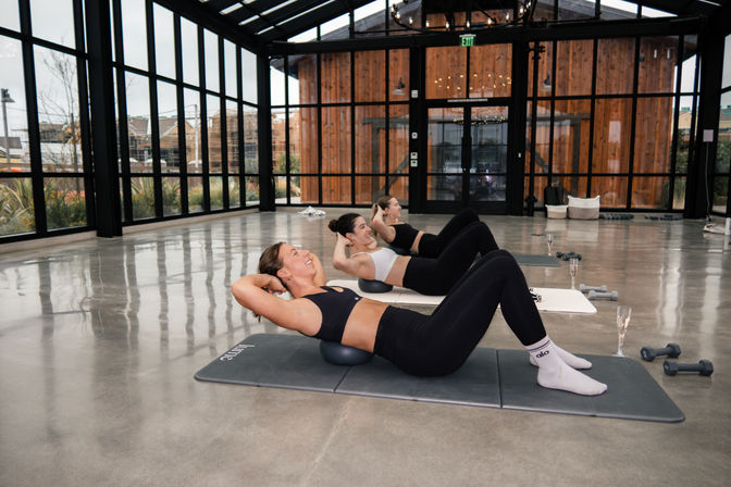Three women in athletic wear doing core crunches on small stability balls in a bright glass-walled fitness studio with concrete floor, mats, dumbbells and champagne flutes.