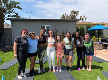 Eight women in athletic wear smiling and posing on yoga mats in a sunny suburban backyard with palm trees, a blue patio umbrella, and exercise mats