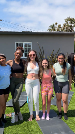 Eight women in athletic wear smiling and posing on yoga mats in a sunny suburban backyard with palm trees, a blue patio umbrella, and exercise mats