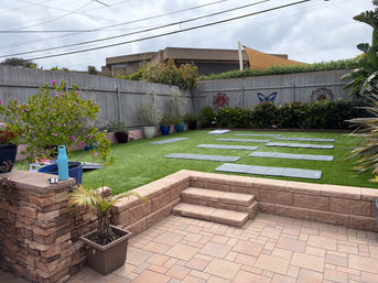Residential backyard patio with stone steps leading to artificial turf arranged with multiple yoga mats, potted plants and a wooden fence decorated with sun and butterfly art under a cloudy sky.