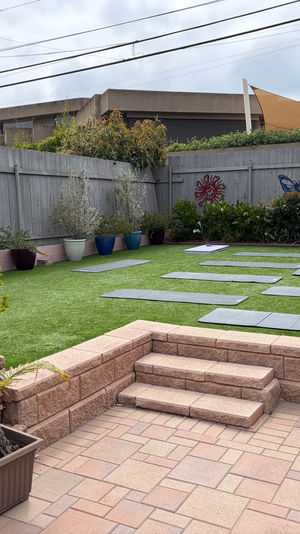 Residential backyard patio with stone steps leading to artificial turf arranged with multiple yoga mats, potted plants and a wooden fence decorated with sun and butterfly art under a cloudy sky.