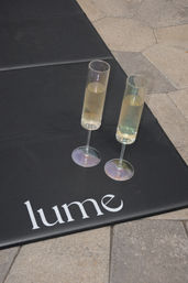 Two slender champagne flutes with pale bubbly resting on a black mat with white lettering, placed on outdoor stone tile pavers.