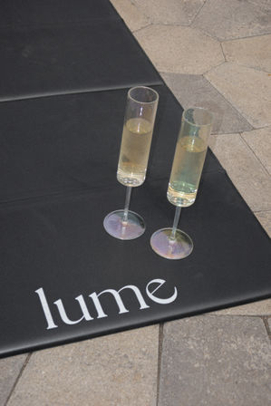 Two slender champagne flutes with pale bubbly resting on a black mat with white lettering, placed on outdoor stone tile pavers.