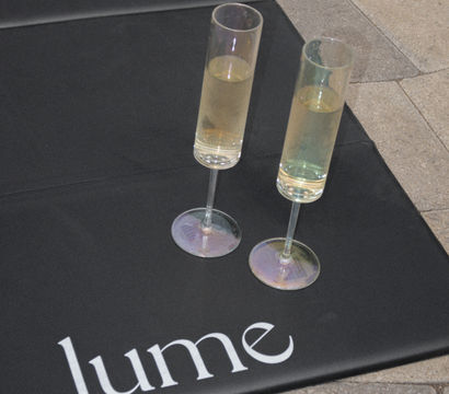 Two slender champagne flutes with pale bubbly resting on a black mat with white lettering, placed on outdoor stone tile pavers.